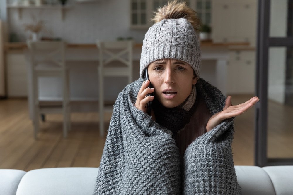 Furnace. Unhappy young woman covered in warm plaid wearing knitted hat, complaining about lack of heating at home in telephone conversation, calling service.