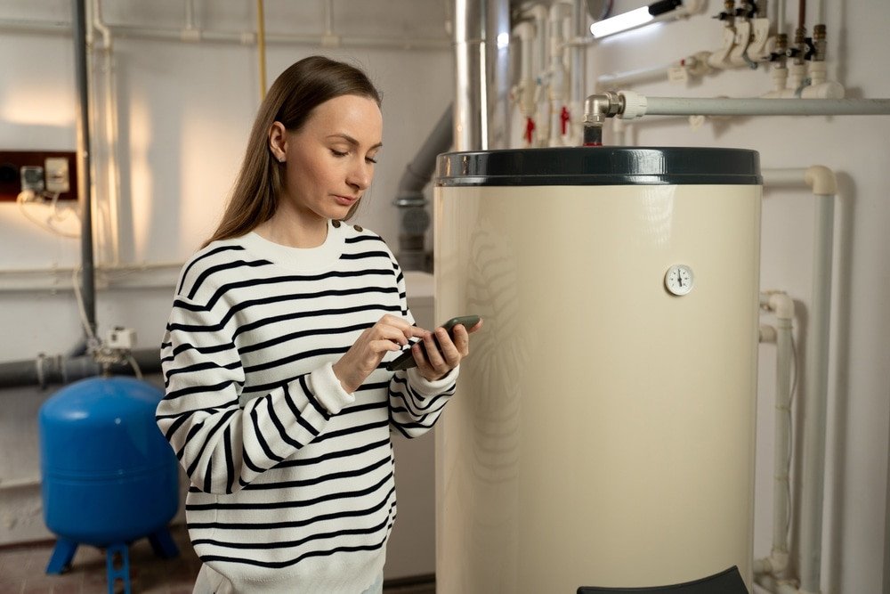 hot water heaters. A young woman examines a non-operational boiler with concern, holding a phone in her hand, likely seeking help or planning to call for repair services. The basement setting indicates a residential.