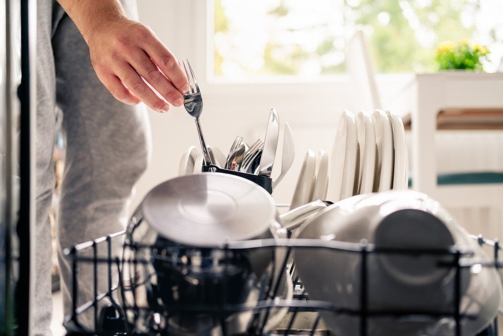 Hard Water. Dish washer machine in kitchen. Man loading dishwasher. Washing plates. Fork in hand. Full of cutlery. Clean or dirty. Pot and tableware. Household chores and housework in family. Unloading utensils.