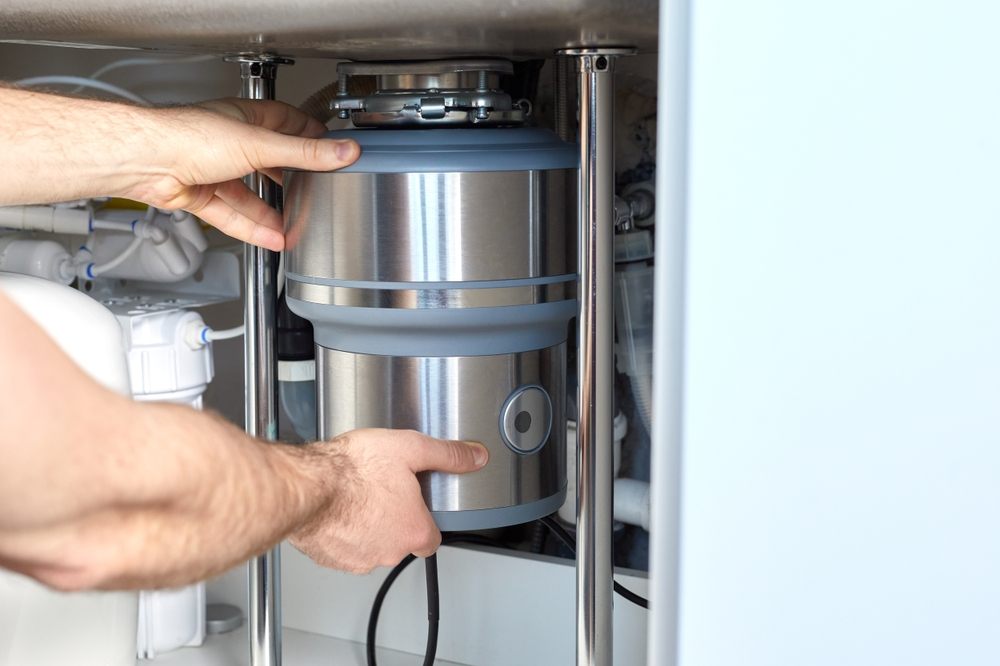 Common Summer Plumbing Problems in Calgary. Clogged Garbage Disposals. A plumber with gloved hands installs a household waste shredder for the kitchen sink. A technician installs or repairs a household waste shredder in the kitchen.
