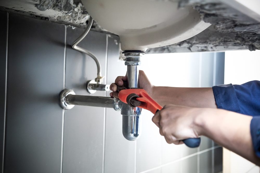 Can Plumbing Services in Calgary Help with Renovation Projects? Bathrooms. A plumber uses a wrench to repair the water pipe under the sink. Maintenance is needed to fix the water leak in the bathroom. With a red wrench, a plumbing installation concept.