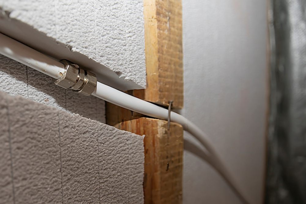 How Do Calgary Plumbers Manage Poly B Pipe Replacements? opening drywall to access plumbing lines. Close‑up of white poly B pipes joined by a metal compression fitting passing through a plastered and wood‑framed wall opening, illustrating precise plumbing installation and construction detail.