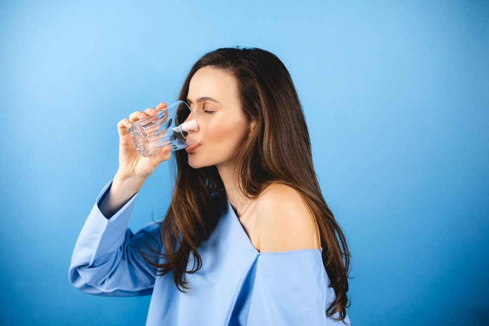 How Can Aquality Improve Your Home's Water Quality? A young woman wearing a blue pullover holds a transparent glass of clear, fresh, pure still water with closed eyes, isolated on a blue background. This is a concept of proper nutrition and healthy fast food choices. 